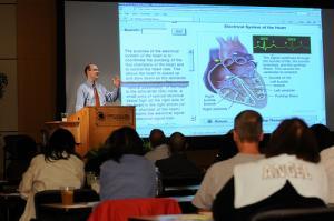 lecturer stands at podium gesturing in front of a screen depicting a heart while students look on